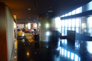 Eine geräumige Business-Lounge am Hong Kong International Airport mit großen Fenstern, hohen Decken mit elektrischem Licht, Sitzgruppen einschließlich Stühlen und Sofas, Tischen, einem Teppichboden und einer zentralen Säule.