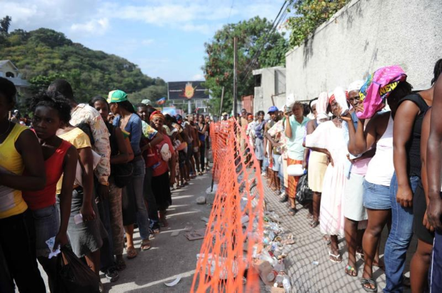 Eine lange Schlange von Menschen wartet vor einem Zaun und einer Wand, um ihre Stimmen in einer Wahllokal abzugeben, mit Bäumen, Gebäuden, Pfählen, Drähten und einer Reklamewand im Hintergrund.