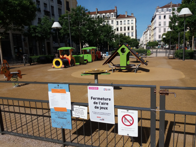 Umzäunter Spielplatz mit Schildern, umgeben von Bäumen, Laternenmasten, Gebäuden und einem sichtbaren Himmel auf einem Stadtplatz.
