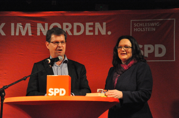 Zwei strahlende Menschen auf einer Bühne vor einem Tisch mit Gegenständen und einem Mikrofon, mit einem Banner im Hintergrund.