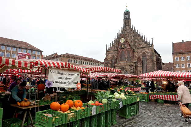 Ein belebter Markt in Nürnberg, Deutschland, mit Obst, Gemüse, Menschen, Zelten und einem Kirchturm im Hintergrund.