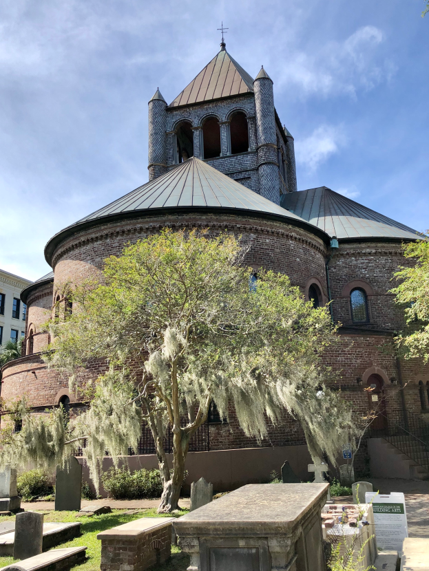 Die Kirche des Heiligen Grabes in Charleston, South Carolina, mit einem Turm und einem Baum davor, umgeben von Gebäuden mit Fenstern, einer Treppe mit Geländer und einem bewölkten Himmel im Hintergrund, mit Gras und einem Schild unten.