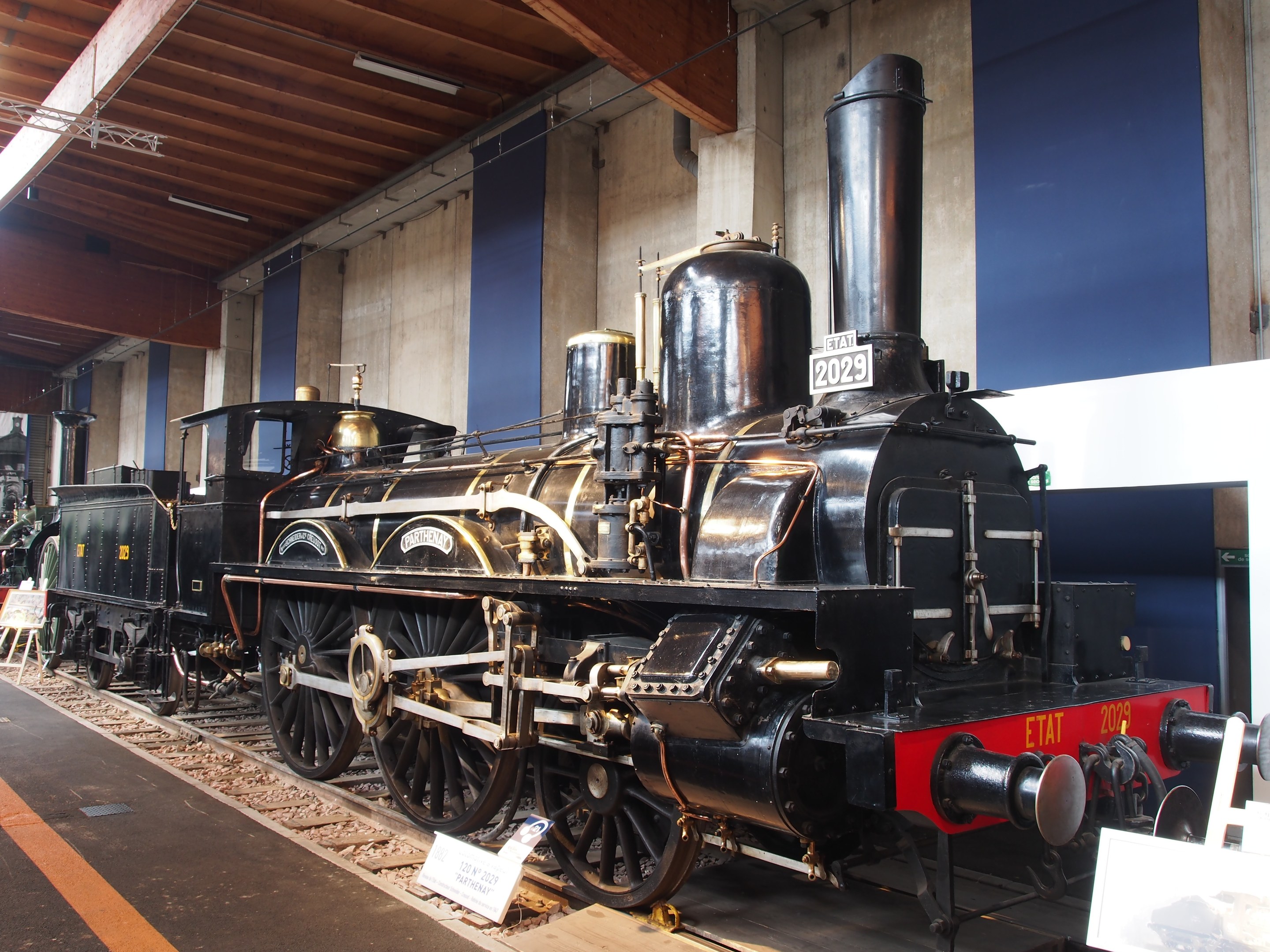 Eine gro├če, alte Dampflokomotive in einem Museums Setting mit umliegenden Eisenbahnschienen, Informationsschildern und anderen Ausstellungen, vor einer Wand mit Deckenbeleuchtung.