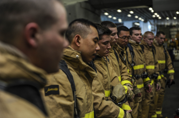 Firefighters in uniform standing in a line, illuminated by background lights.