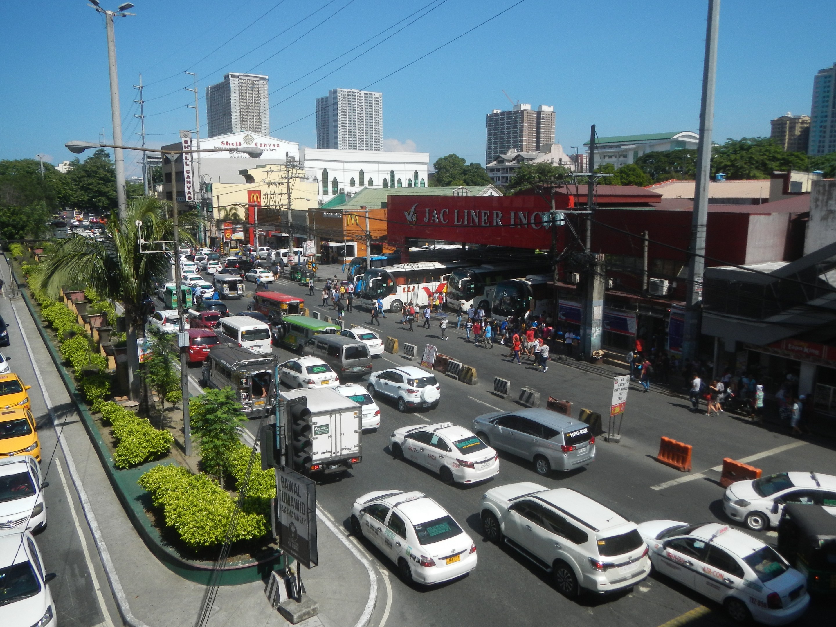 Eine belebte Stadtstraße mit Autos, Bussen, Fußgängern, Grünflächen, Straßenlaternen, Strommasten und Gebäuden unter einem klaren blauen Himmel.