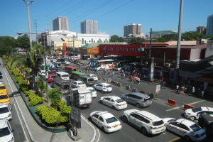 Eine belebte Stadtstraße mit Autos, Bussen, Fußgängern, Grünflächen, Straßenlaternen, Strommasten und Gebäuden unter einem klaren blauen Himmel.
