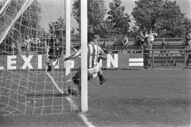 Ein Schwarz-Weiß-Foto eines Fußballspielers, der den Ball ins Tor schießt, mit einem Netz auf der linken Seite und einer Gruppe von Menschen auf den Tribünen im Hintergrund, mit Bäumen und einem klaren Himmel.