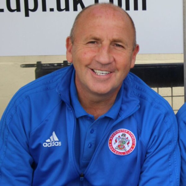Zwei Männer in einem Raum, einer in einem blauen Jackett, der neben einem anderen Mann lächelt, mit einer Wandtafel im Hintergrund.