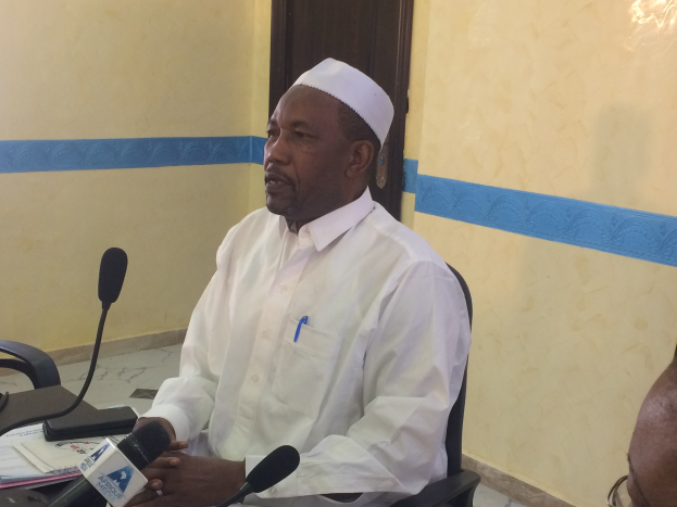 Ein Mann in einem weißen Kleid und einer Mütze sitzt an einem Tisch mit Mikrofonen, spricht, während Papiere und Gegenstände auf dem Tisch sichtbar sind, mit einem anderen Mann mit Brille rechts von ihm, in einem Raum mit einer Wand und einer Tür im Hintergrund.