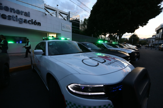 Eine Reihe von Polizeiwagen vor einem Gebäude mit Text geparkt, umgeben von Menschen, Bäumen, Laternenmasten, Drähten und einem bewölkten Himmel.