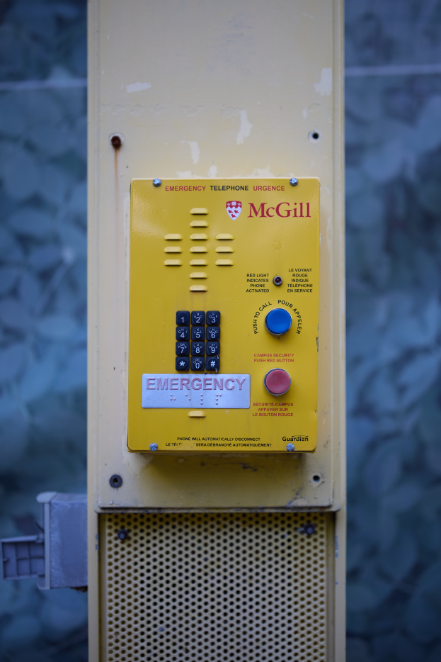 Ein gelber Notrufhörer an einer Gebäudeseite montiert, mit Tasten und Text zum Absetzen von Notrufen, vor einer Wand.