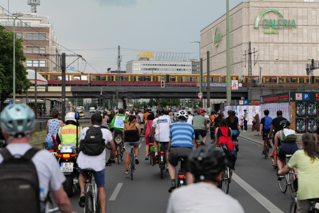 Eine Gruppe von Menschen, die auf Fahrrädern eine Straße mit hohen Gebäuden entlangfahren, einige tragen Helme und Taschen, mit einem Zug auf einem Eisenbahn Gleis, Strommasten, Bäumen und einem klaren blauen Himmel im Hintergrund.
