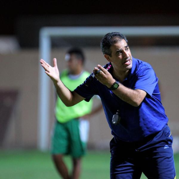 Ein Mann in einem blauen T-Shirt und Hose steht auf einem Fußballfeld und hält ein Mikrofon in der Hand, während er auf Spieler zeigt, und im Hintergrund ist eine Person in einem grünen Kleid und ein Torpfosten zu sehen.