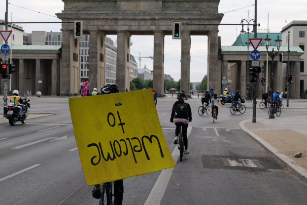 Eine Gruppe von Menschen auf Fahrrädern am Brandenburger Tor in Berlin vorbei, einer hält ein gelbes Schild, mit Laternen, Verkehrszeichen, Gebäuden und Bäumen im Hintergrund unter einem klaren blauen Himmel.