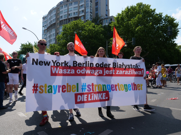 Eine Gruppe von Menschen, die eine Straße in Berlin, Deutschland, entlanggehen und ein Banner mit der Aufschrift "Stay Rebel Stronger Together" tragen, mit Bäumen, Laternenpfählen und Gebäuden an der Straße und einem bewölkten Himmel.