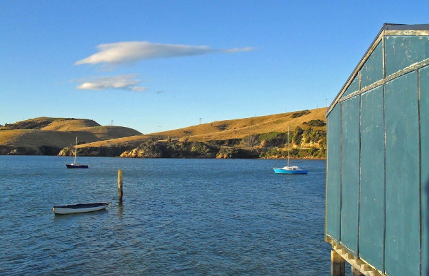 Boote und Pfähle im Wasser, ein hölzernes Haus auf der rechten Seite, Bäume und Berge mit Windmühlen im Hintergrund und Wolken am Himmel.
