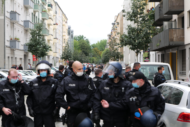 Eine Gruppe von Polizisten in Masken und Helmen geht eine Straße entlang, die von parkenden Autos gesäumt ist, flankiert von Gebäuden mit Balkonen und Geländern, mit Bäumen und einem klaren Himmel im Hintergrund.