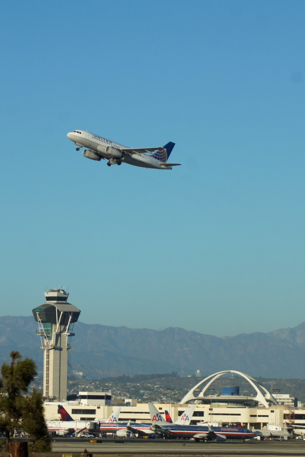 Ein Flughafenblick mit einem weißen Kontrollturm, parkenden Flugzeugen, entfernten Gebäuden und einem Flugzeug am Himmel.