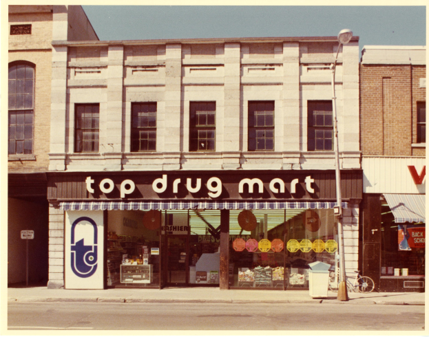 Top Drug Mart Ladengeschäft an einer Stra√enecke mit einem Gebäude, einer Stra√enlampe, einem abgestellten Fahrrad, einem Müllcontainer und einem bewölktem Himmel.