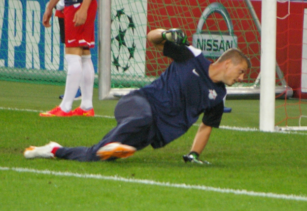 Ein Fußballer mit Handschuhen taucht auf, um einen Ball vor einem Tor zu fangen, mit zwei Personen, einem Netz und einem Banner mit Text im Hintergrund.