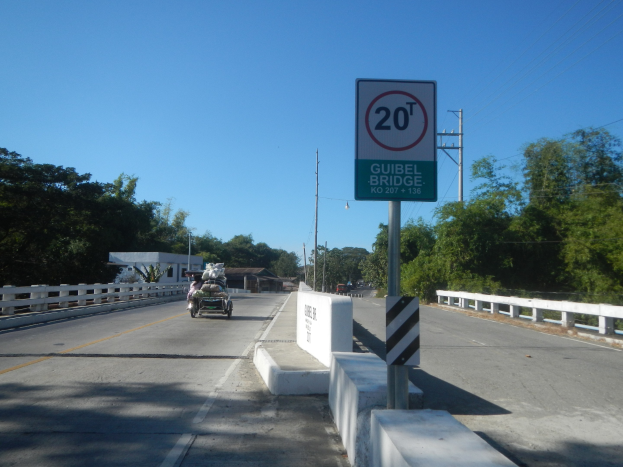Ein Auto, das auf einer Brücke mit einem Tempolimit-Schild, Metallgeländern auf beiden Seiten, Bäumen im Hintergrund und Himmel darüber fährt.