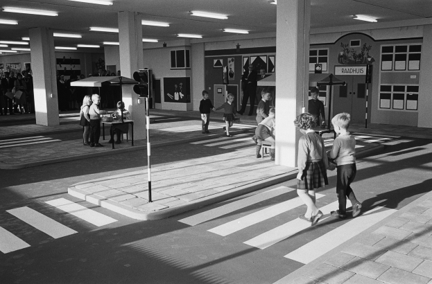 Schwarz-weißes Foto von Kindern, die eine Straße öberqueren, mit Menschen in der Nähe und einem Verkehrszeichen im Vordergrund.