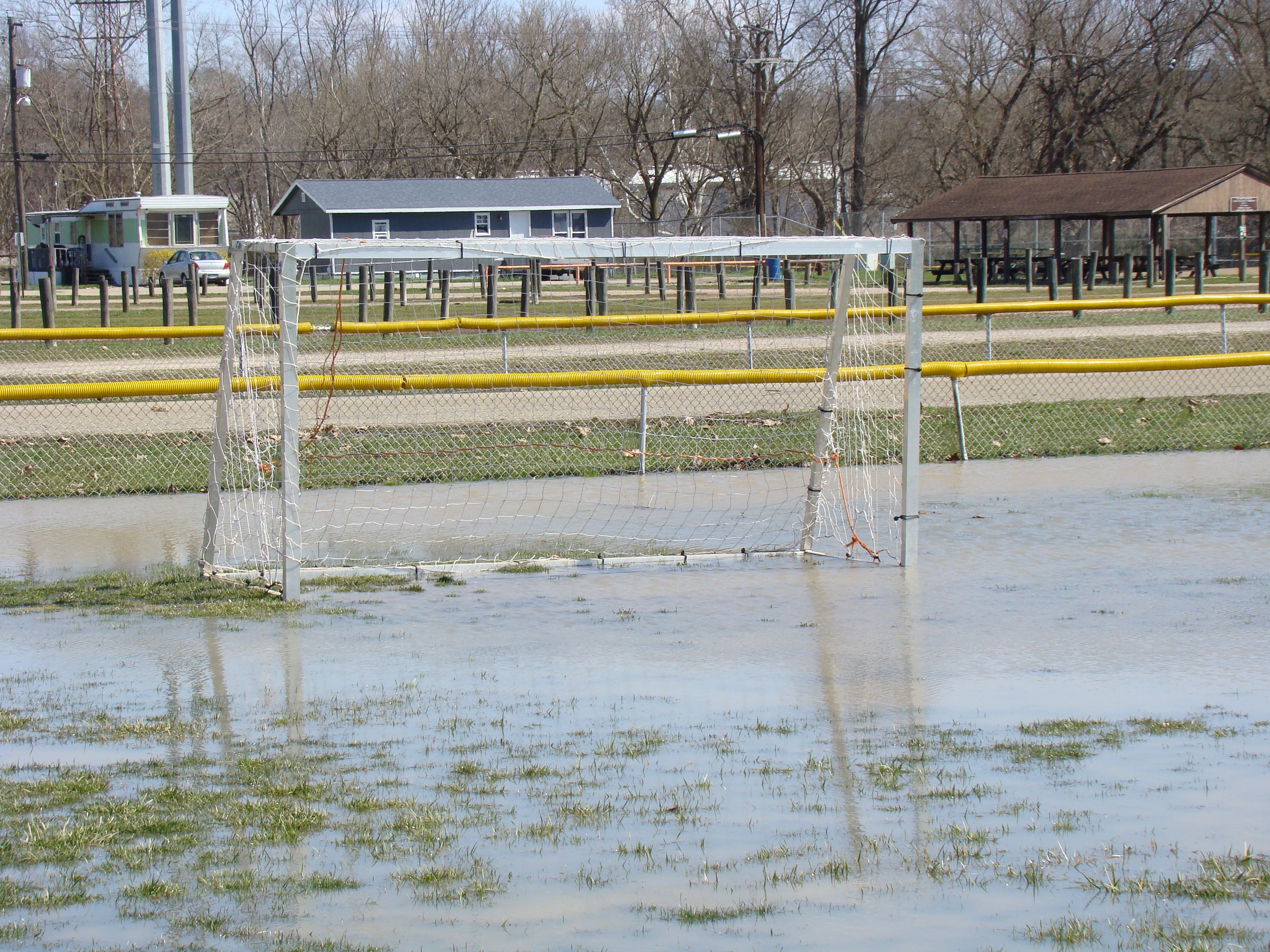 Ein Fußballtor steht in einem überfluteten Feld mit umgebenem Gras und Wasser, mit Hütten, Pfählen, Bäumen, Fahrzeugen und einem klaren blauen Himmel im Hintergrund.
