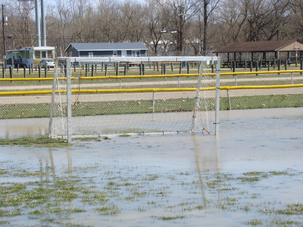Ein Fußballtor steht in einem überfluteten Feld mit umgebenem Gras und Wasser, mit Hütten, Pfählen, Bäumen, Fahrzeugen und einem klaren blauen Himmel im Hintergrund.