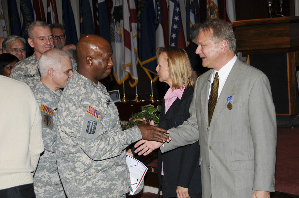 Eine Gruppe von Menschen, die mit zwei Personen Handschlag gibt, Flaggen im Hintergrund, ein Podium mit Mikrofon und eine Topfpflanze in der Nähe.