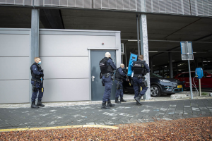 Eine Gruppe von Polizeibeamten steht vor einem Gebäudeeingang in einer Parkanlage, umgeben von Fahrzeugen, Pfählen, Brettern und verstreuten Steinen.