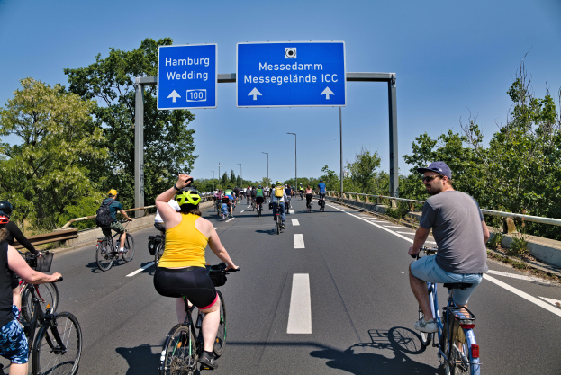 Eine Gruppe von Radfahrern in Helmen fährt eine Straße mit einer Begrenzung auf einer Seite und Bäumen auf der anderen entlang, unter einem klaren blauen Himmel mit Laternen im Hintergrund und einem Schild oben, das eine Radtour in Hamburg anzeigt.