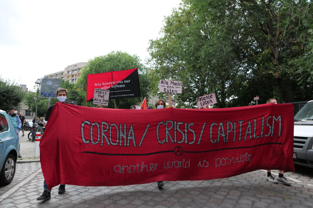 Eine Gruppe von Menschen marschiert mit einer roten Fahne, auf der 'Corona-Krise/Kapitalismus' steht, eine Straße entlang, mit parkenden Fahrzeugen auf beiden Seiten und Gebäuden, Bäumen und einem klaren blauen Himmel im Hintergrund.