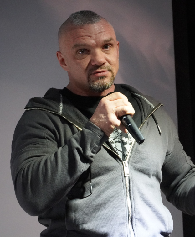 Comedian in einem schwarzen Anzug und wei├čem Hemd, der ein Mikrofon halt, mit zurückgek├Ąmmtem Haar und einem selbstbewussten Ausdruck, vor einer Wand stehend.