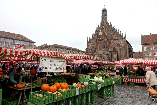 Ein belebter Markt in Nürnberg, Deutschland, mit verschiedenen Früchten und Gemüsen auf dem Stand, Menschen beim Einkaufen, Zelte und Gebäude mit einem Uhrenturm im Hintergrund bei klarem Himmel.
