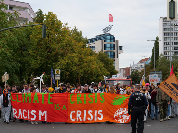 Eine Gruppe von Menschen marschiert auf einer von Bäumen gesäumten Straße und hält ein Banner mit der Aufschrift "Klima-Krise ist eine Krise", mit Gebäuden und einem klaren blauen Himmel im Hintergrund.