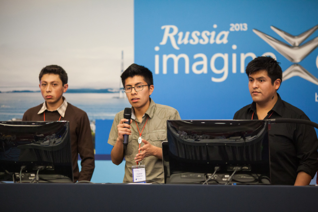 Drei Jungen stehen neben einem Schreibtisch mit zwei Computersystemen, einer spricht in ein Mikrofon und trägt eine ID-Karte mit rotem Anhänger, vor einer Wand mit einer Werbetafel, auf der 'Russia Imagine 2013' steht.