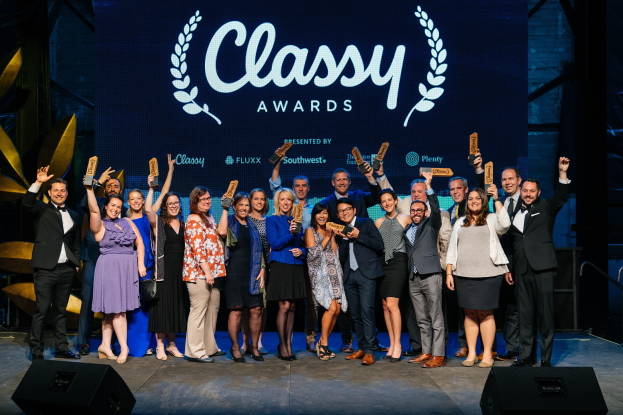 Eine Gruppe von Menschen auf einer Bühne, die lächeln und Auszeichnungen halten, mit Lautsprechern darunter und einem Hintergrundbild mit "Classy Awards 2019".