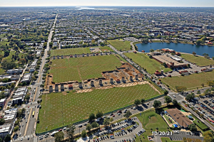Luftaufnahme einer Stadt mit Gebäuden, Bäumen, Gras, Straßen mit Fahrzeugen, Wasser und bewölktem Himmel, die einen zentralen großen Feld als Parkplatz zeigt.
