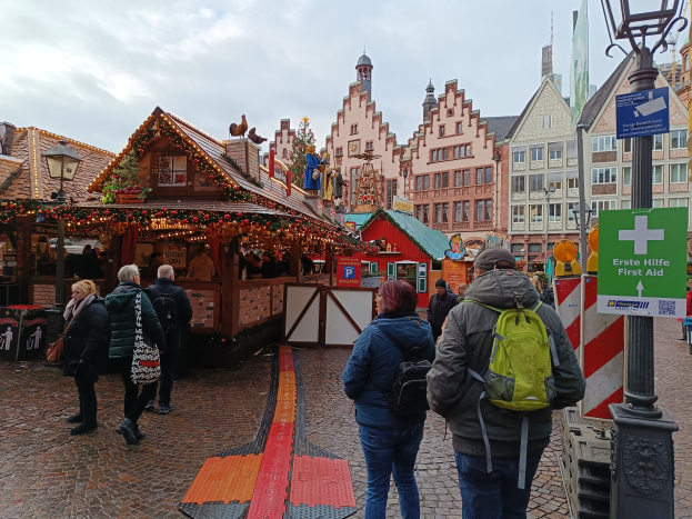 Eine Gruppe von Menschen geht eine Kopfsteinpflasterstraße entlang neben einem Weihnachtsmarkt in Nürnberg, Deutschland, wobei einige Taschen, Laternenmasten und Texttafeln zu sehen sind und Gebäude mit Fenstern und ein bewölkter Himmel im Hintergrund.