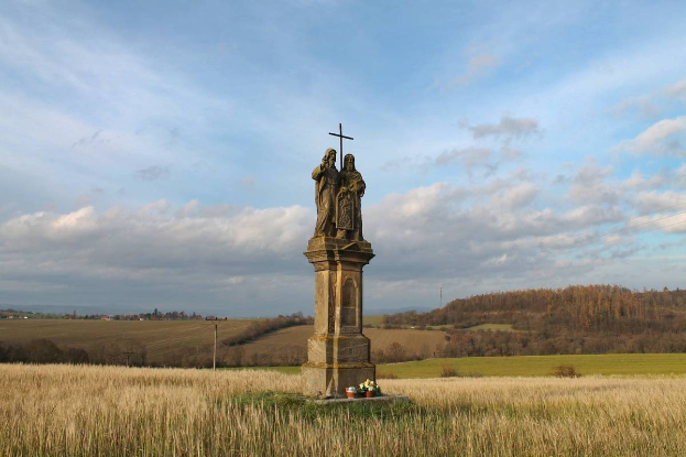 Ein Denkmal mit einem Kreuz darauf steht auf einem Feld umgeben von Pflanzen und Blumen, mit Bäumen, Pfählen und einem bewölkten Himmel im Hintergrund, als Gedenkstätte für Holocaust-Opfer in Deutschland.