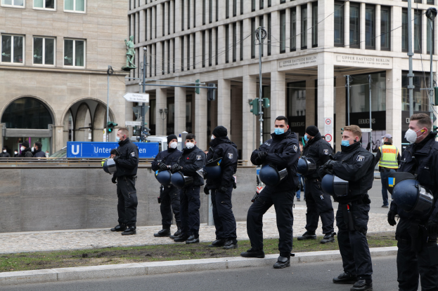 Eine Gruppe von Polizisten in schwarzen Uniformen und Masken steht vor einem Berliner Gebäude mit Glasfenstern und Säulen, einige halten Helme, mit Laternen, Verkehrsampeln, Texttafeln, einer Statue und anderen Menschen im Hintergrund auf einem grasbewachsenen Boden.