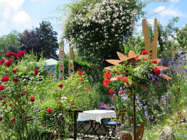 Ein Garten mit bunten Blumen und Pflanzen, der einen Tisch mit einer Decke und Stühlen zeigt, vor Bäumen und einem bewölkten Himmel an der Chelsea Flower Show.