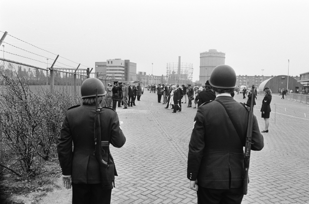 Zwei Polizisten in Helmen und mit Waffen in den Händen stehen nebeneinander auf einem Bürgersteig, mit einem Zaun und Pflanzen zu ihrer Linken und Gebäuden, Pfählen und einem klaren Himmel im Hintergrund.