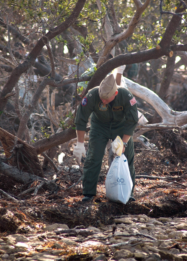 Ein Mann mit Mütze und Handschuhen hält eine Folienabdeckung, während er Müll in einem bewaldeten Gebiet mit Steinen und trockenen Blättern auf dem Boden sammelt.
