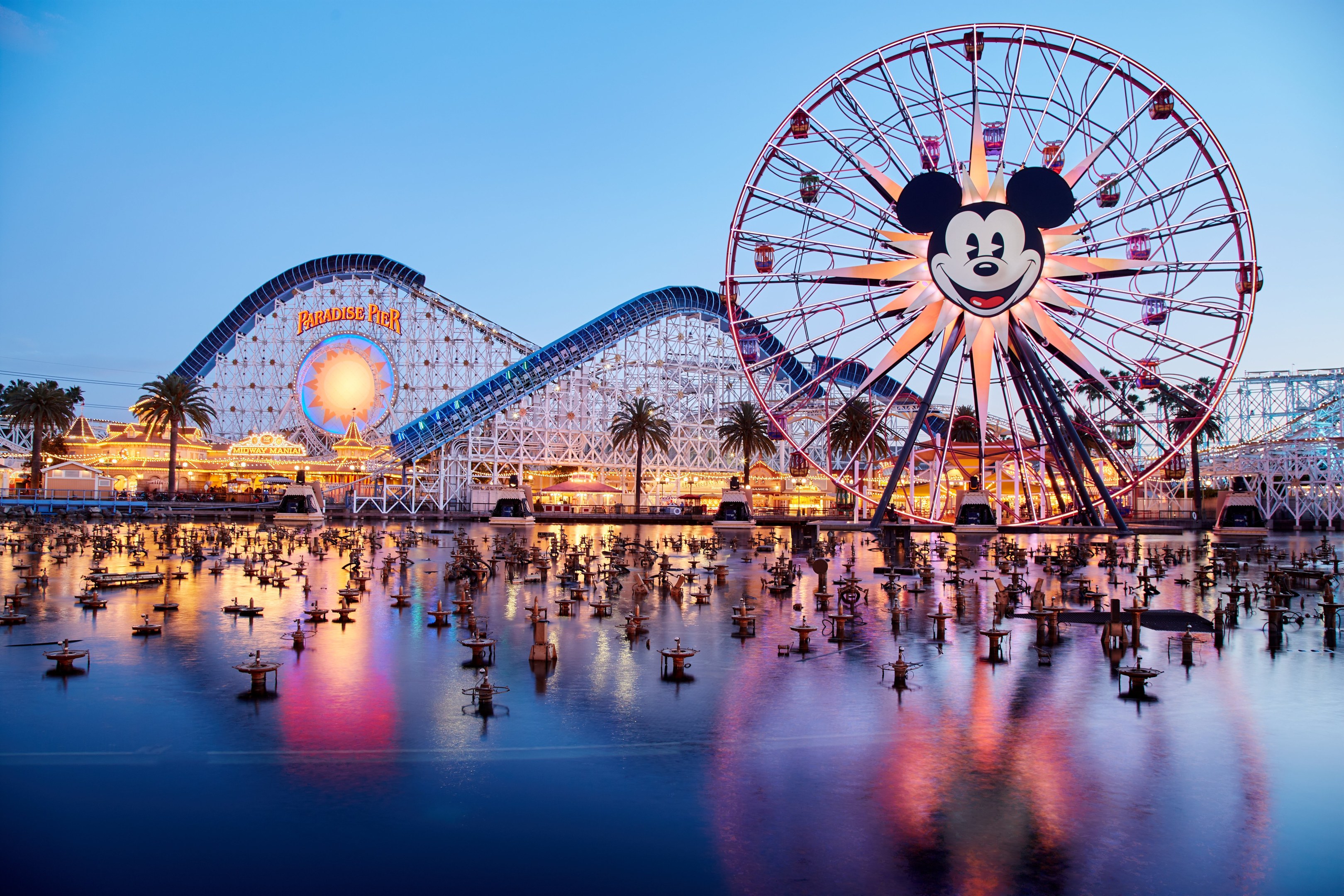 Eine lebendige Dämmerungsszene im Disneyland California Adventure mit einem riesigen Mickey-Maus-Rad im Vordergrund, einer Achterbahn im Hintergrund und Booten auf dem Wasser, mit einem Himmel aus Orangen, Pinks und Purpurs und silhouettenartigen Bäumen.