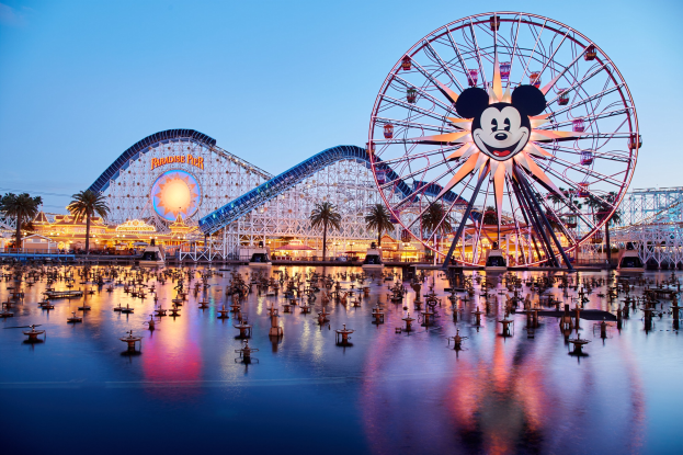 Eine lebendige Dämmerungsszene im Disneyland California Adventure mit einem riesigen Mickey-Maus-Rad im Vordergrund, einer Achterbahn im Hintergrund und Booten auf dem Wasser, mit einem Himmel aus Orangen, Pinks und Purpurs und silhouettenartigen Bäumen.