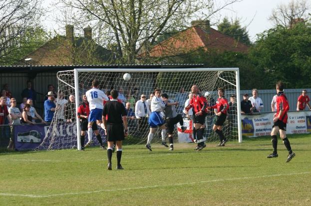 Fußballspieler spielen auf einem Feld mit einem Tornetz, während Zuschauer dahinter stehen; im Hintergrund sind Bäume und Häuser zu sehen.