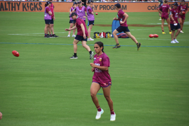 Gruppe von Frauen, die Australian Rules Football auf einem grünen Feld spielen, mit verstreuten Bällen und einem Banner im Hintergrund, einige tragen Mützen und Turnschuhe.