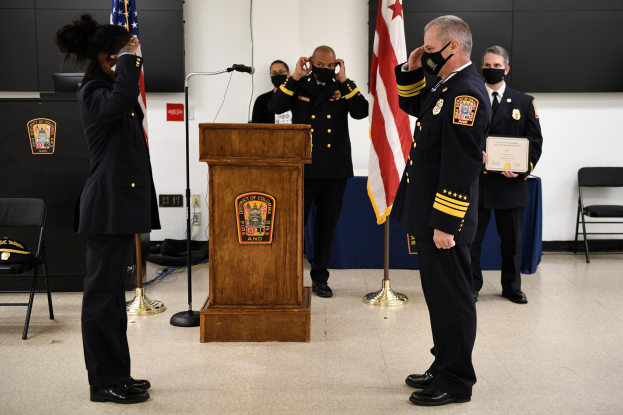 Gruppe uniformierter Personen in Masken vor einem Rednerpult bei einer Vereidigung eines neuen Feuerwehrchefs, der eine Urkunde hält, mit Flaggen und Sitzplätzen im Hintergrund.