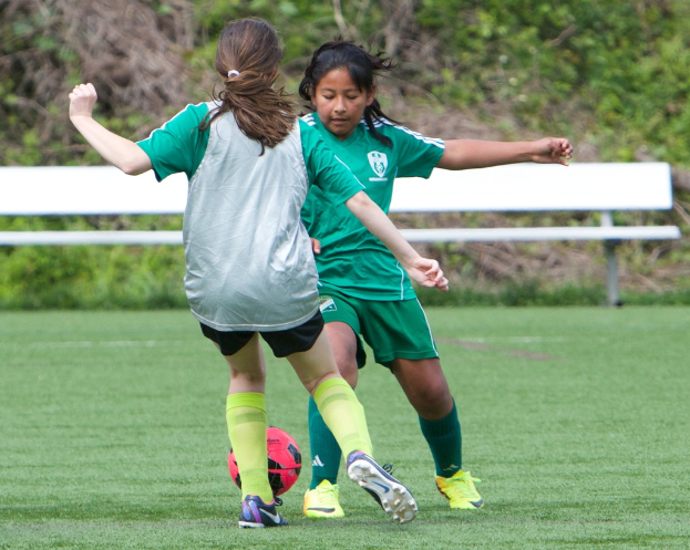 Zwei Mädchen in grünen Trikots spielen Fußball auf einem grünen Rasenfeld mit einer weißen Bank, Bäumen und Pflanzen im Hintergrund und einer Wand unten.